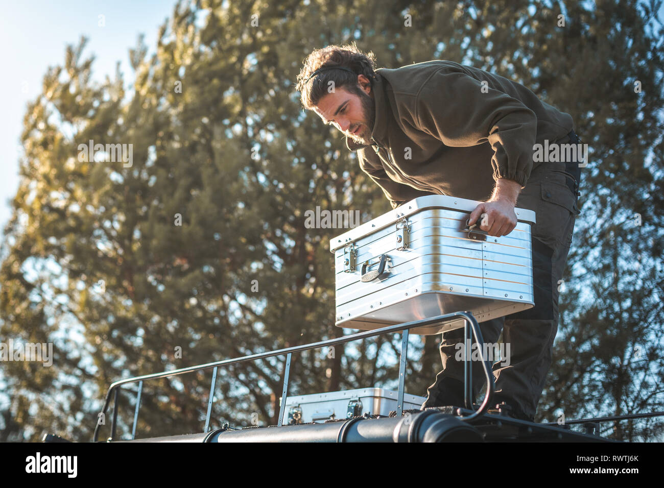 A man is handling a metal box on the roof of a camper van surrounded by nature Stock Photo