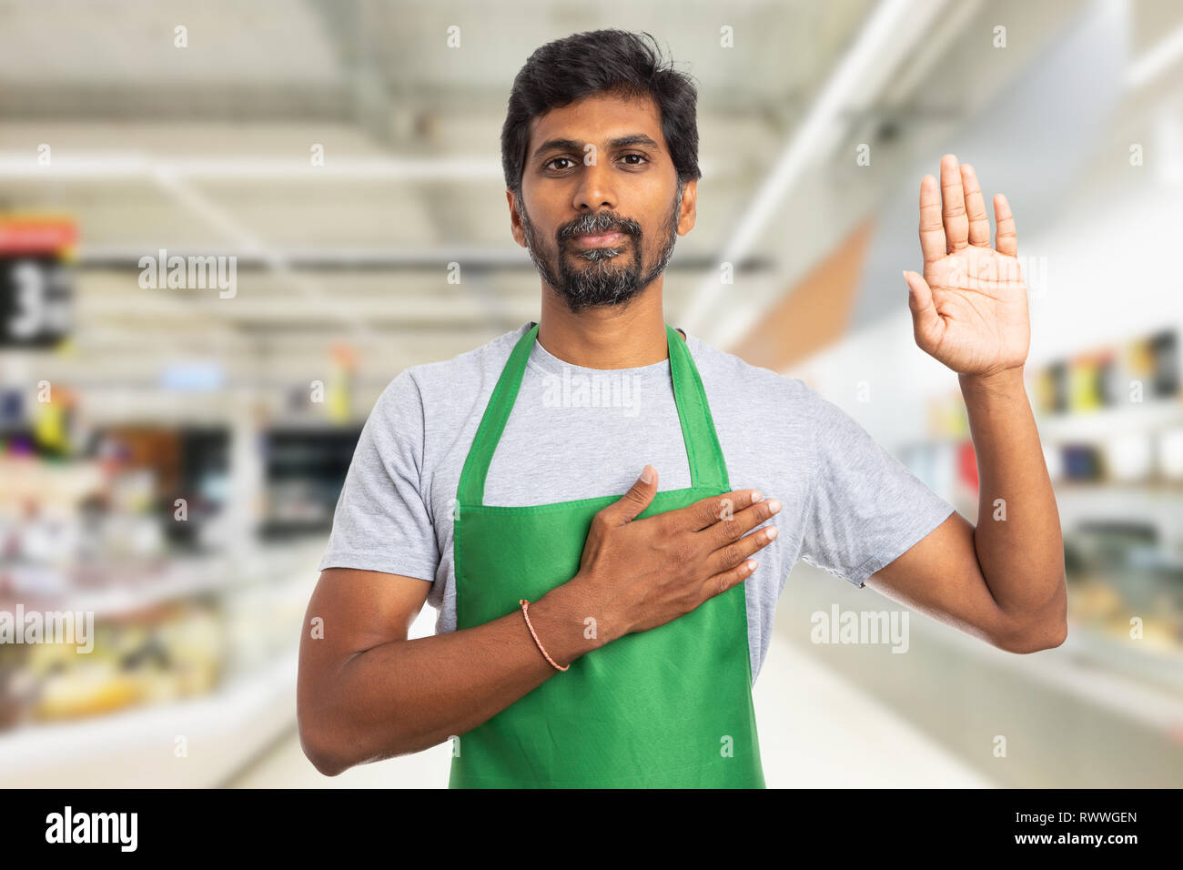 Serious trustworthy indian male hypermarket or supermarket employee making honest oath gesture with hand on heart and palm up Stock Photo