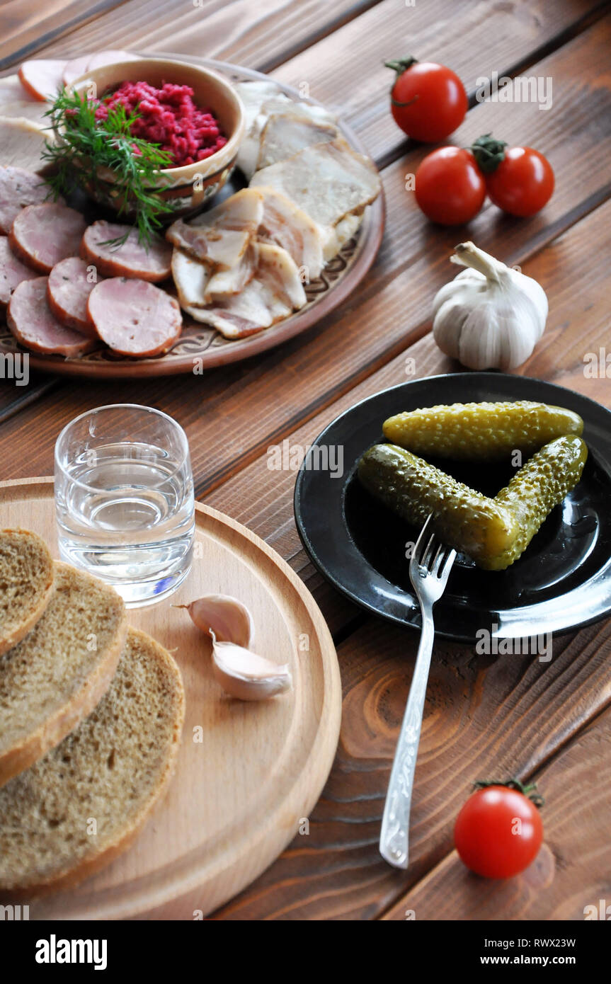 A glass of vodka with pickles, rye bread, fresh tomatoes, garlic, lard, sausage and grated horseradish on the wooden table, selective focus Stock Photo