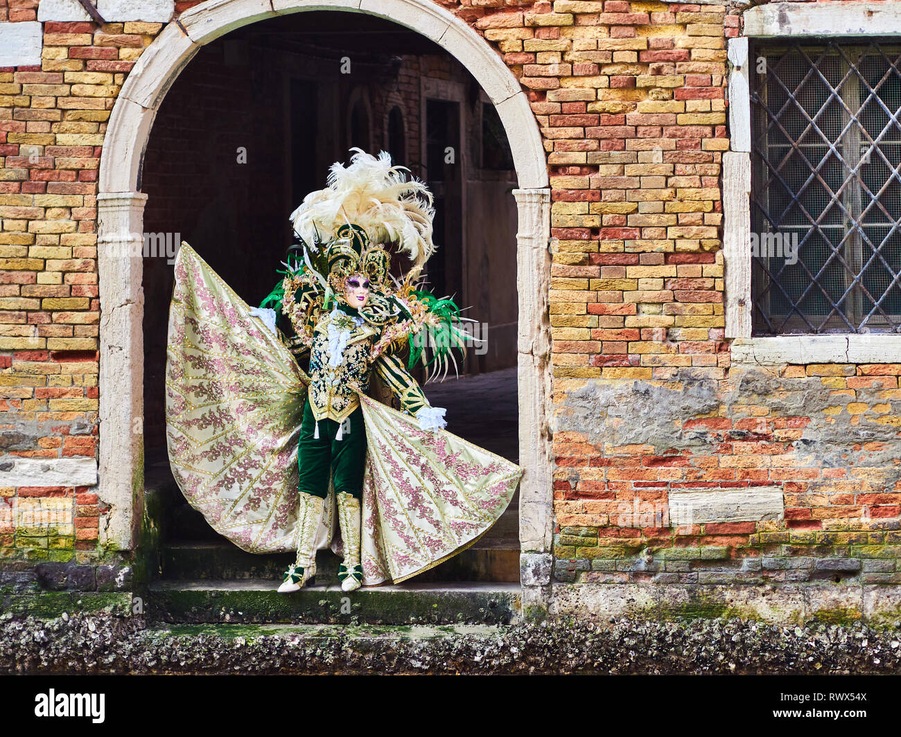Venice, Italy - March 2, 2019 Person dressed in a typical Venetian Costume models under a arch towards a water channel in Venice during the Carnival o Stock Photo