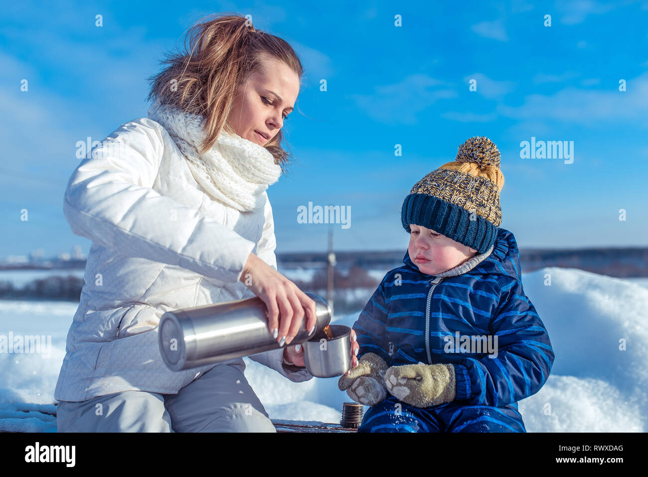 Mom and little boy son 3-4 years old bench winter street, snow snowdrifts in background. Pours into a mug. Thermos with hot tea soup drink, breakfast Stock Photo