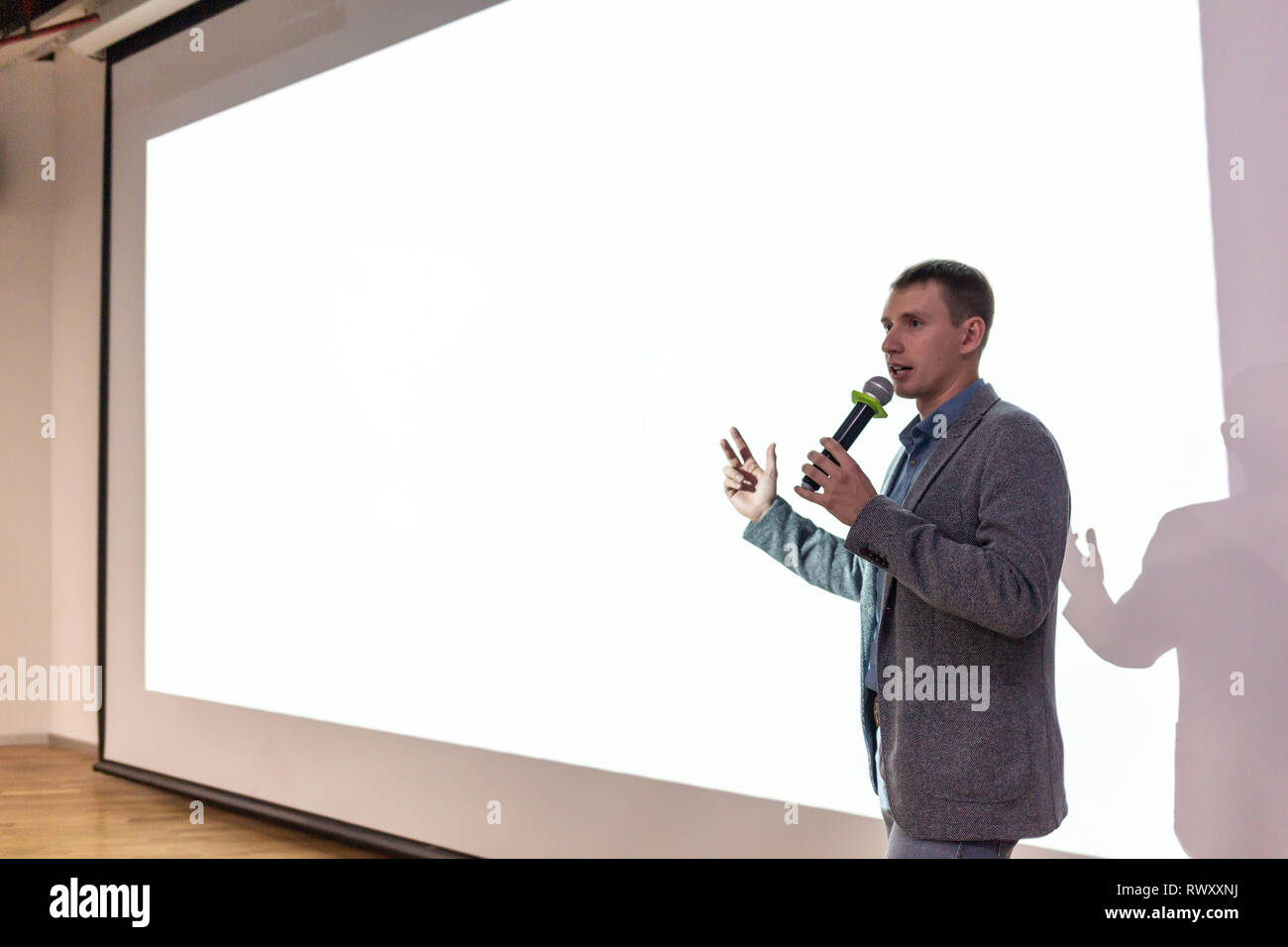 A man near the white screen of the projector with a micraphone talks about the project. Stock Photo