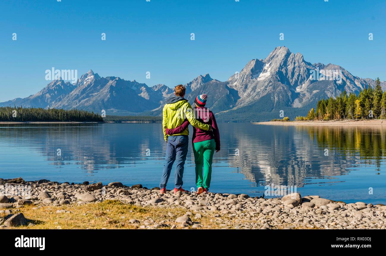 Young woman and young man standing side by side on the shore, looking into the landscape, mountains reflected in the lake Stock Photo