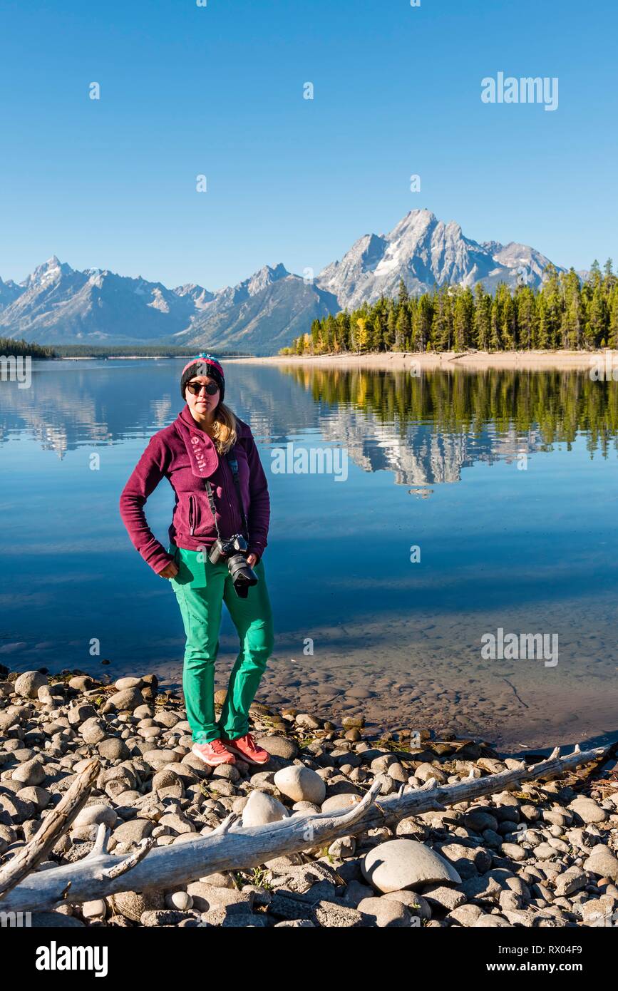 Young woman standing on the shore looking into the camera, mountains reflected in the lake, Colter Bay, Jackson Lake Stock Photo
