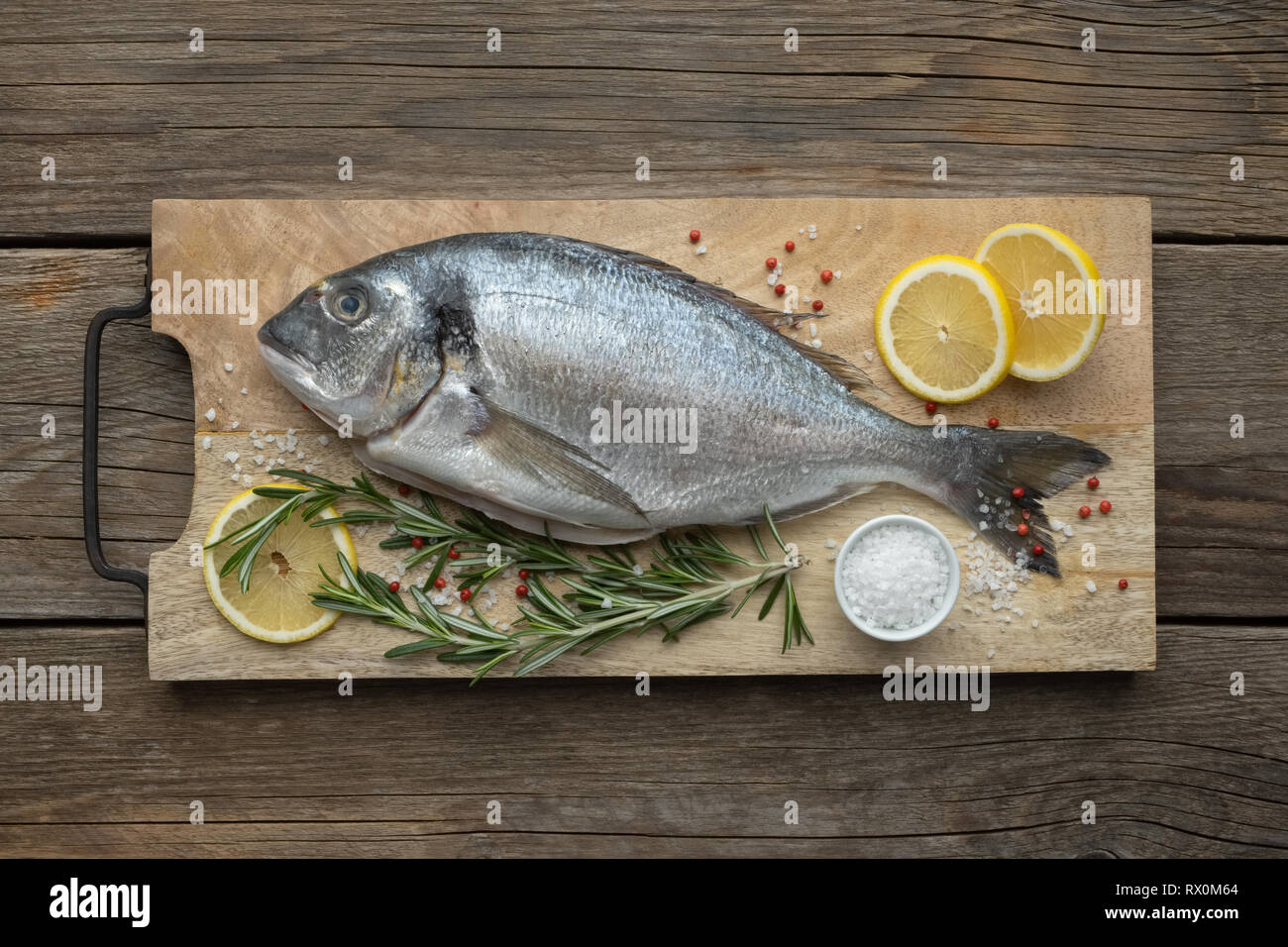 Fresh dorado fish with lemon slices, salt and rosemary on wooden cutting board. Top view. Stock Photo