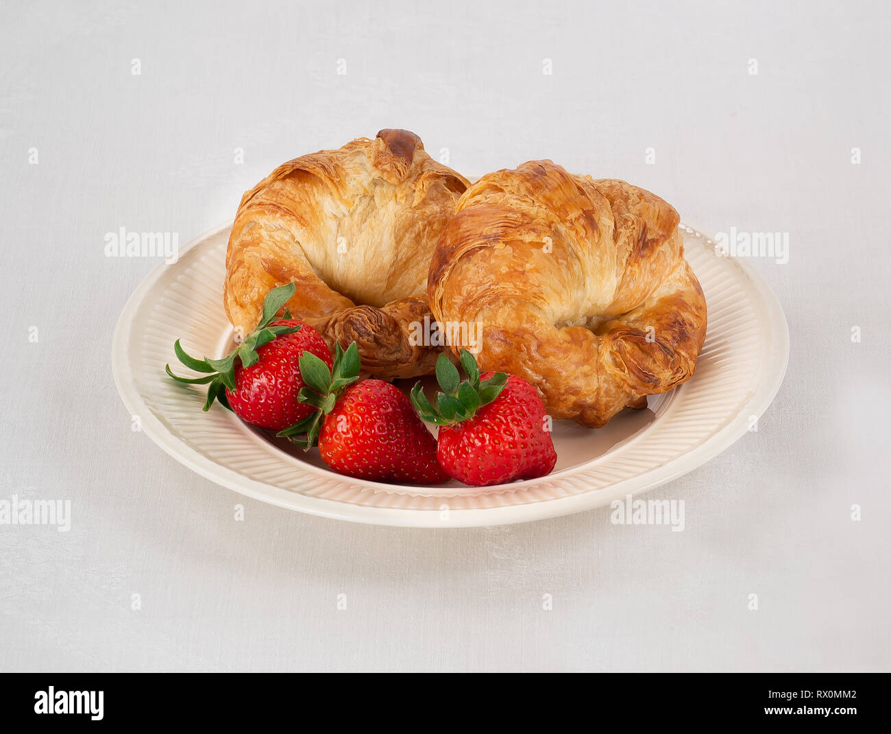 Cream Colored Plate with Two Croissants and Three Fresh Strawberries on a White Linen Tablecloth. Stock Photo