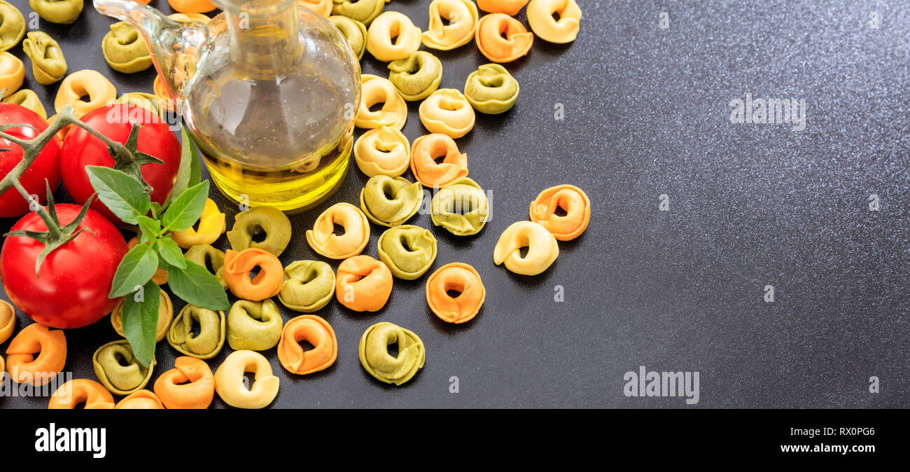 Italian food preparation. Colorful tortellini pasta, tomatoes, olive oil and basil on black background, banner, copy space Stock Photo