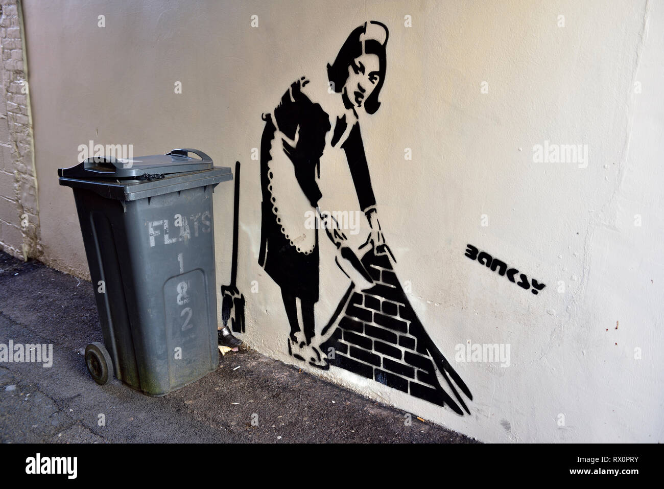 Stencil painting on wall of woman with dustpan with real rubbish bin next to painting, Question if Bansky Stock Photo