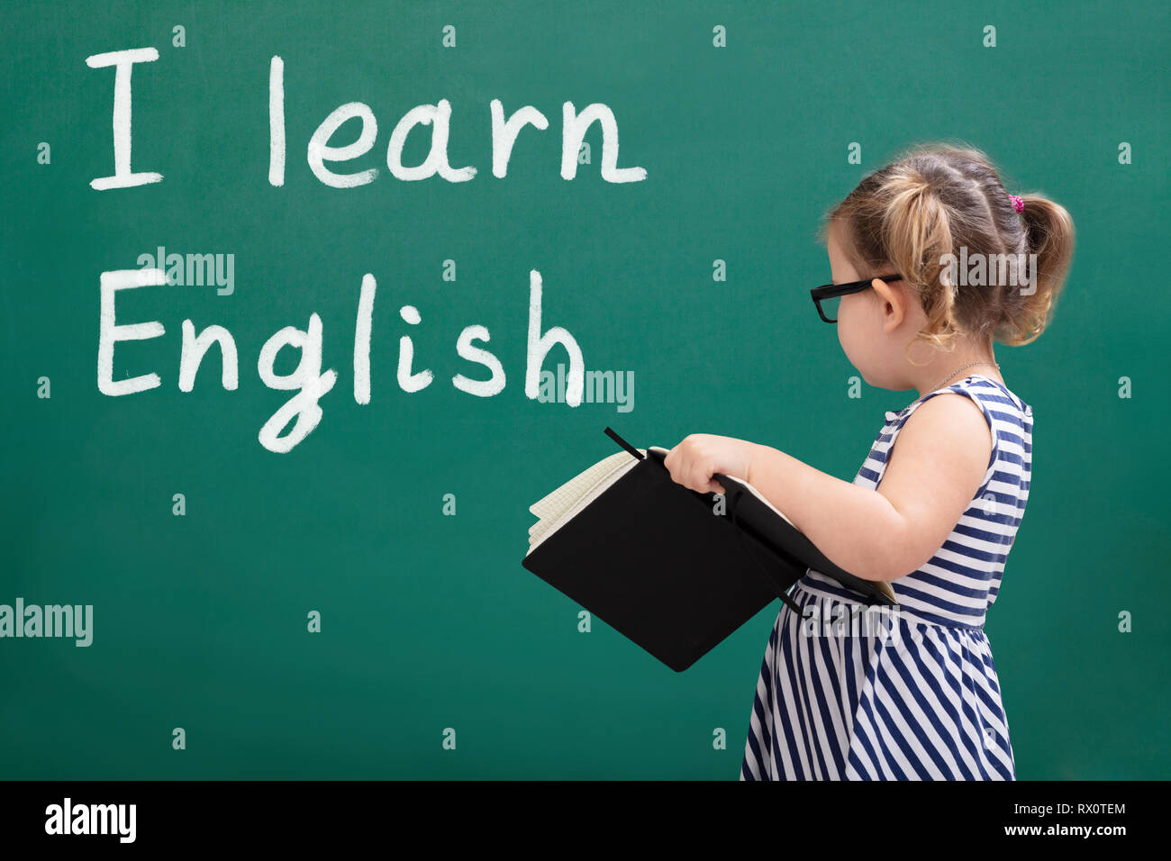 Side View Of Cute Girl Holding A Book With Text Learn English On The Chalkboard Stock Photo