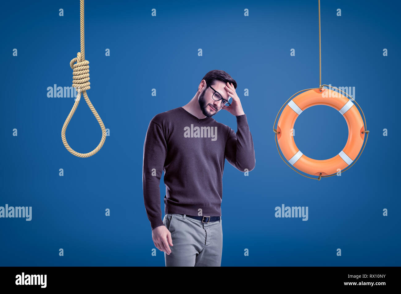 Man in casual outfit standing half-turn with hand on forehead as if choosing between lifebuoy hanging on rope and hangman's noose. Stock Photo