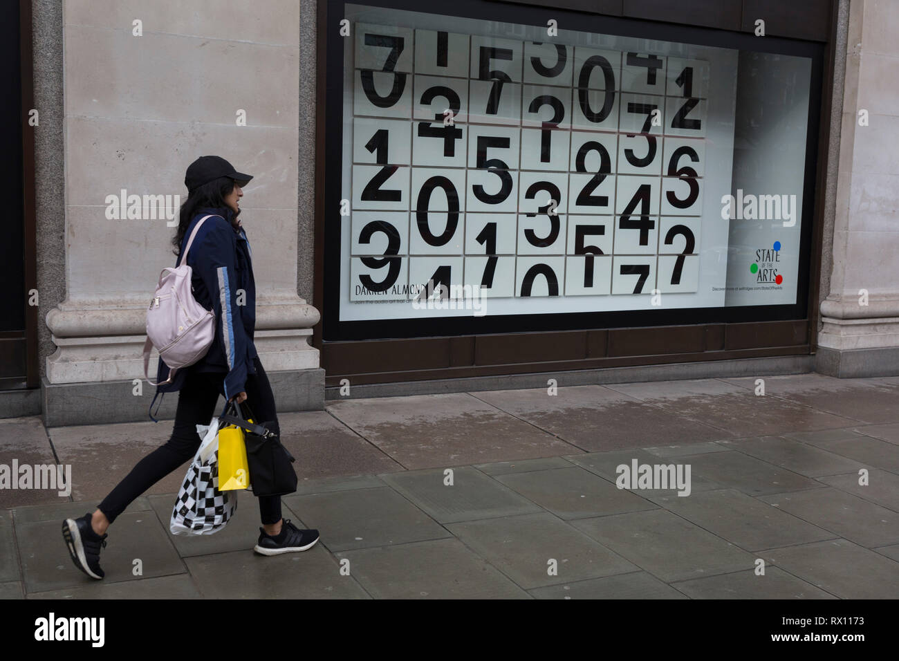 A shopper walks past a window display that features numbers - part of a design theme called 'State of the Arts', at the Selfridges department store on Oxford Street, on 4th March 2019, in London England. Darren Almond's piece ‘Chance Encounter 004’, consists of a grid formed from rectangular panels, featuring fragmented numbers that appear to scroll across the surface.  State of the Arts is a gallery of works by nine crtically-acclaimed artists in Selfridges windows to celebrate the power of public art. Stock Photo