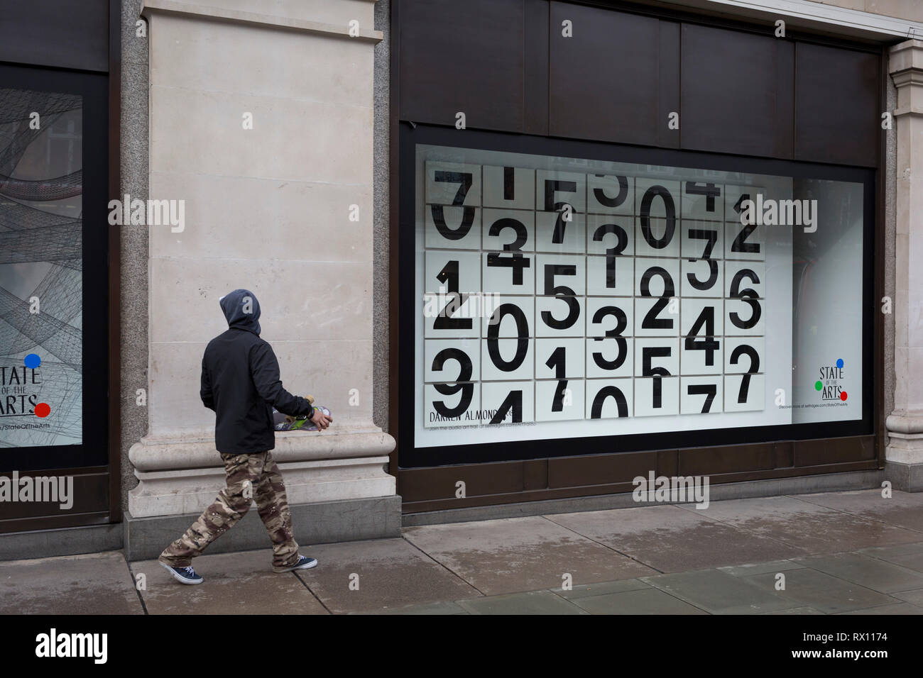 A skateboarder walks past a window display that features numbers - part of a design theme called 'State of the Arts', at the Selfridges department store on Oxford Street, on 4th March 2019, in London England. Darren Almond's piece ‘Chance Encounter 004’, consists of a grid formed from rectangular panels, featuring fragmented numbers that appear to scroll across the surface. State of the Arts is a gallery of works by nine crtically-acclaimed artists in Selfridges windows to celebrate the power of public art. Stock Photo