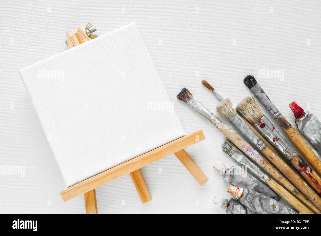 Canvas on easel, paint tubes and bundle of brushes for painting on white canvas background. Top view. Flat lay. Copy space for text. Stock Photo