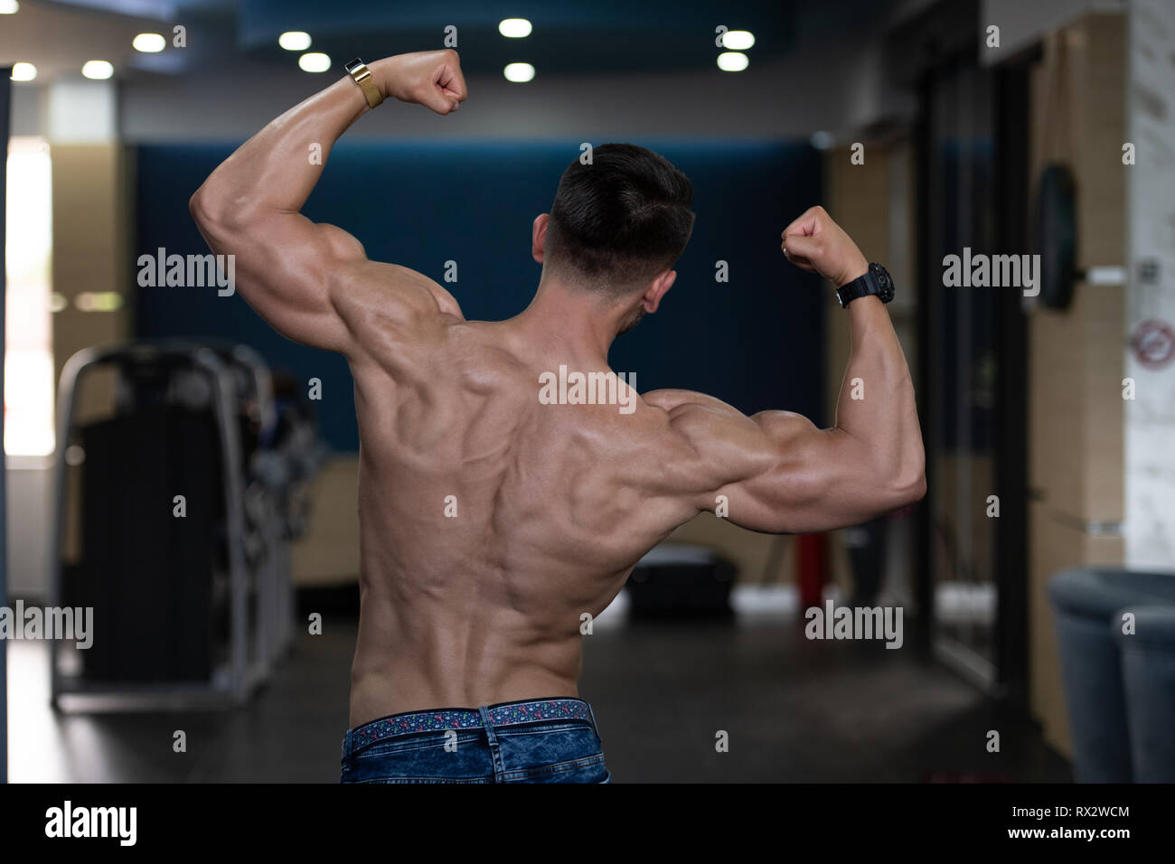 Handsome Young Man In Jeans Standing Strong In The Gym And Flexing Muscles - Muscular Athletic Bodybuilder Fitness Model Posing After Exercises Stock Photo