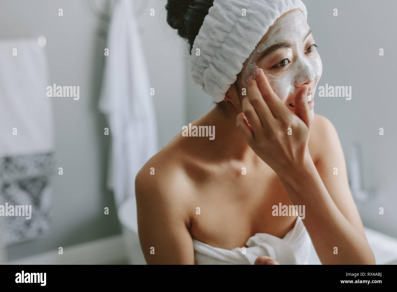 Young asian woman in bathroom applying facial mask on her face. Female in putting face pack onto her face. Stock Photo