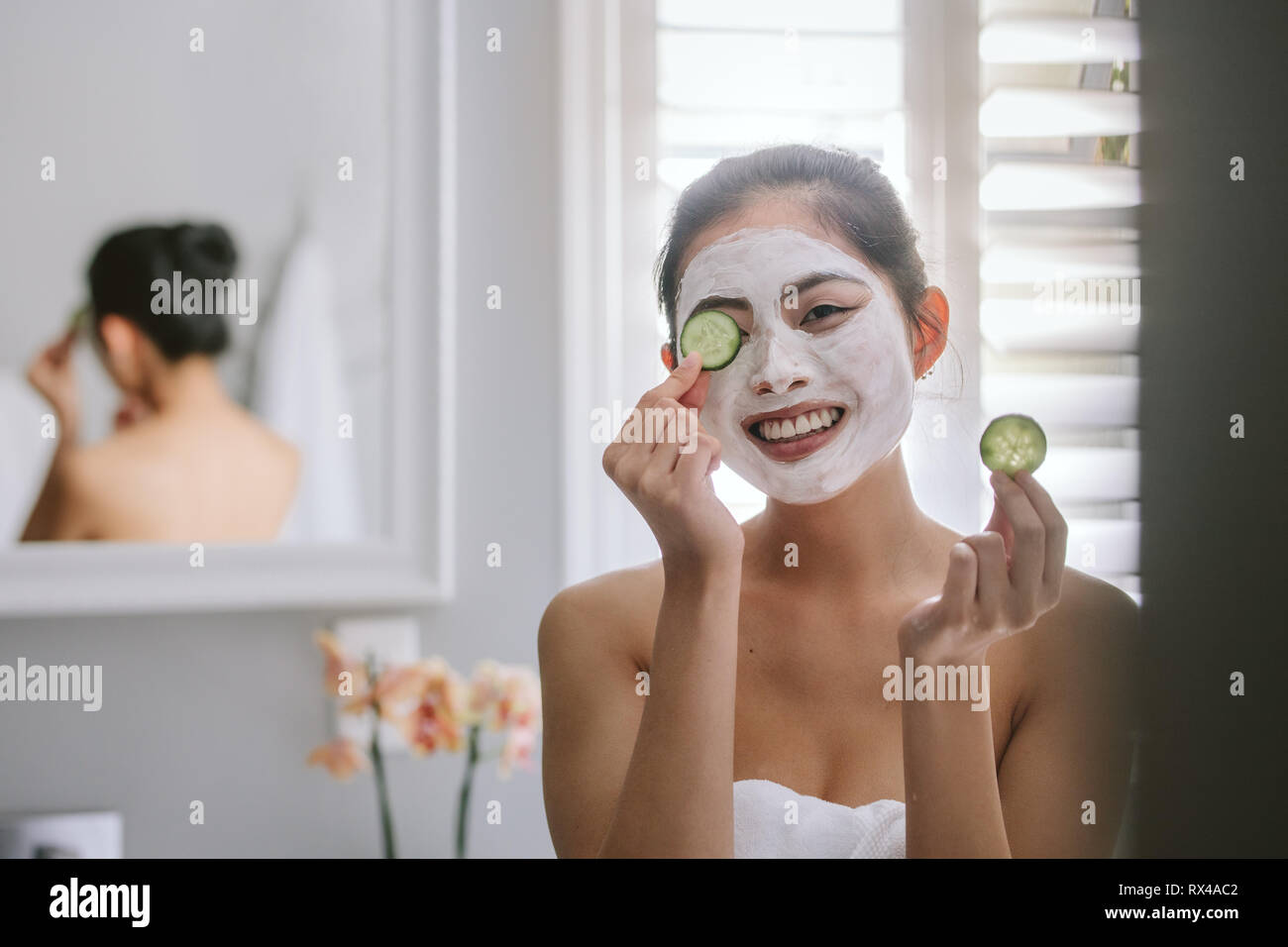 Smiling young woman with face pack holding slice of cucumber to eyes. Happy woman in bathroom having spa facial treatment in morning. Stock Photo
