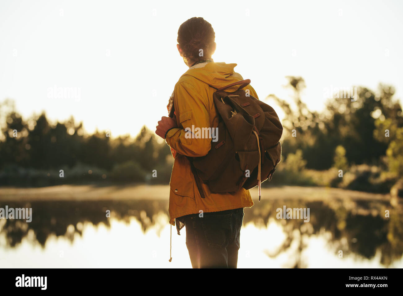 Man wearing a backpack standing near a lake and looking at the evening sun. Tourist exploring nature standing outdoors near a lake with sun in the bac Stock Photo