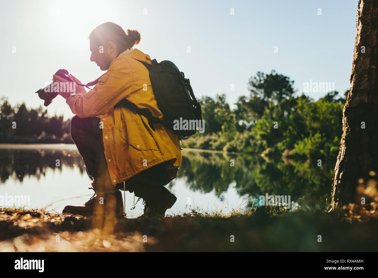 Side view of a traveler adjusting his digital camera sitting beside a lake. Man looking at his dslr camera while taking photos outdoors with sun in th Stock Photo