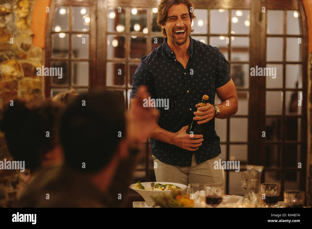 Laughing man opening champagne with friends sitting around table during party. Millennials having a dinner party. Stock Photo