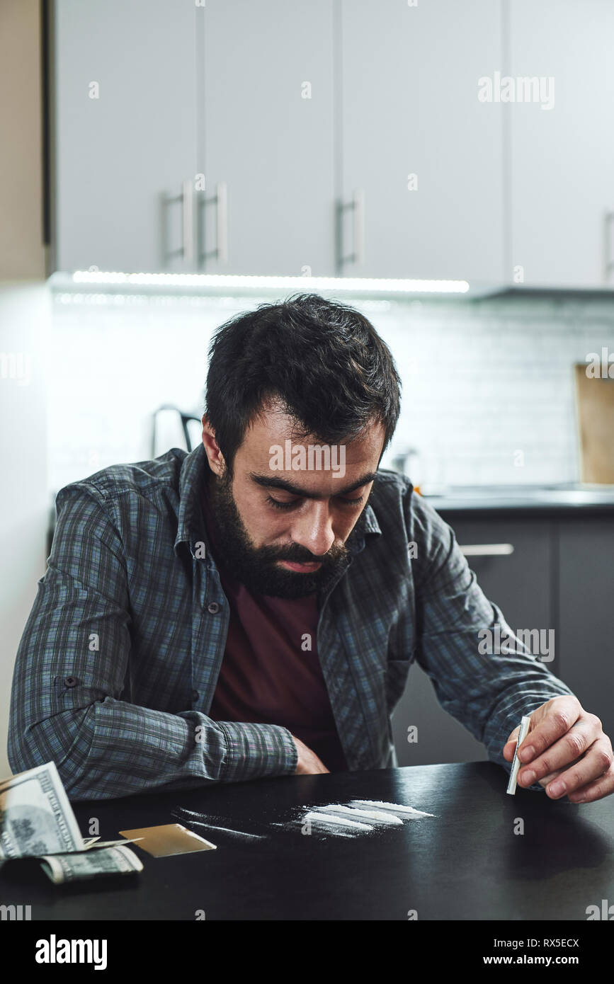 Depressed bearded man sits at the table, preparing to snort a line of cocaine with a rolled up bank note. He is suffering from problems at work and tr Stock Photo