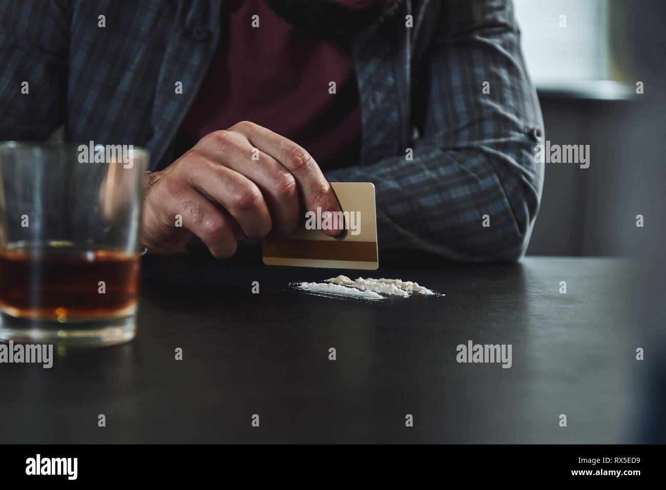 Depressed man sits at the table, preparing to snort a line of cocaine. He is suffering from problems at work and troubles in relationships. A glass of Stock Photo