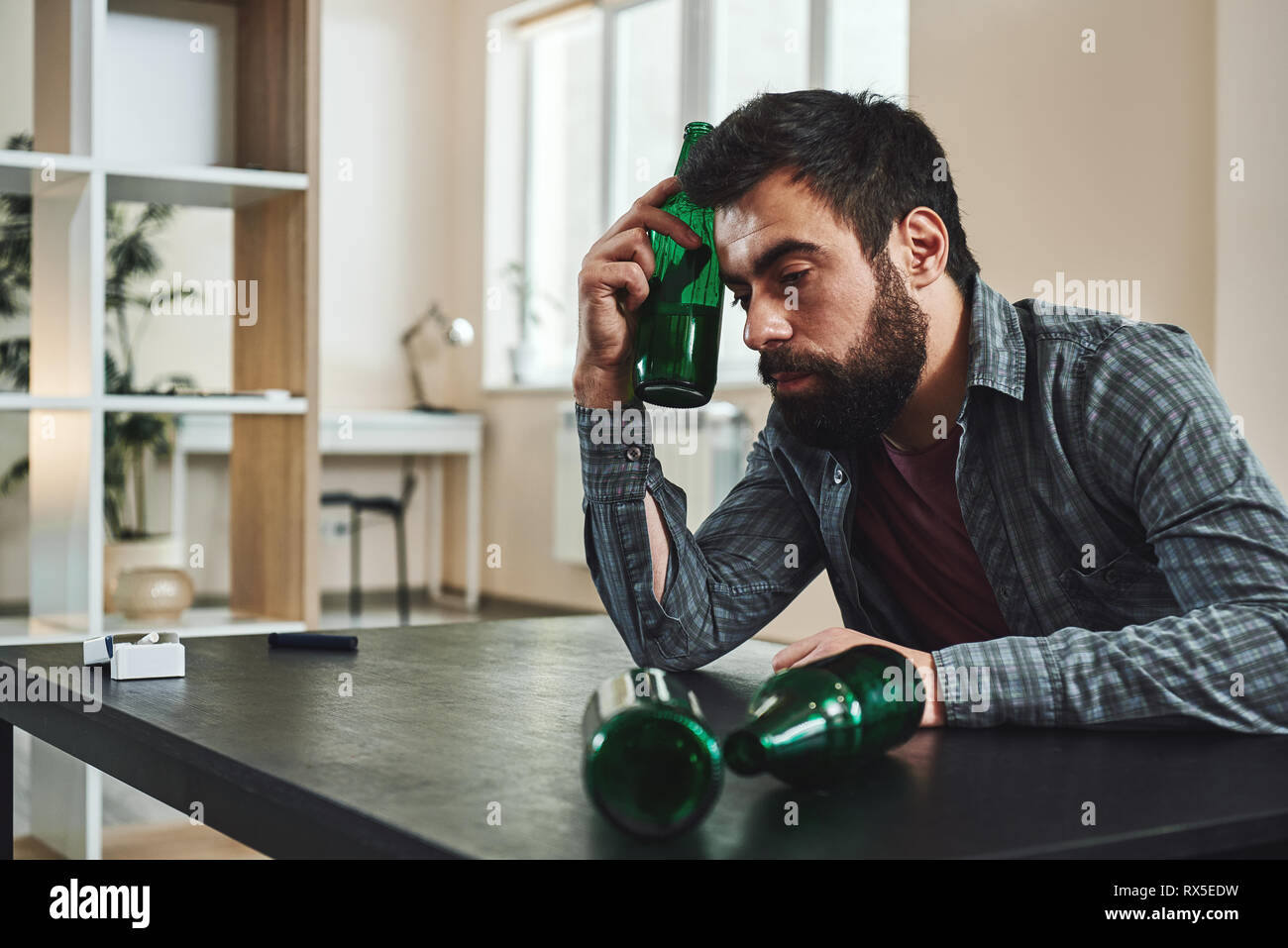 Drinking trouble. Depressed bearded man sits at the table with beer bottle in his hand. He is thinking about problems at work and troubles in relation Stock Photo