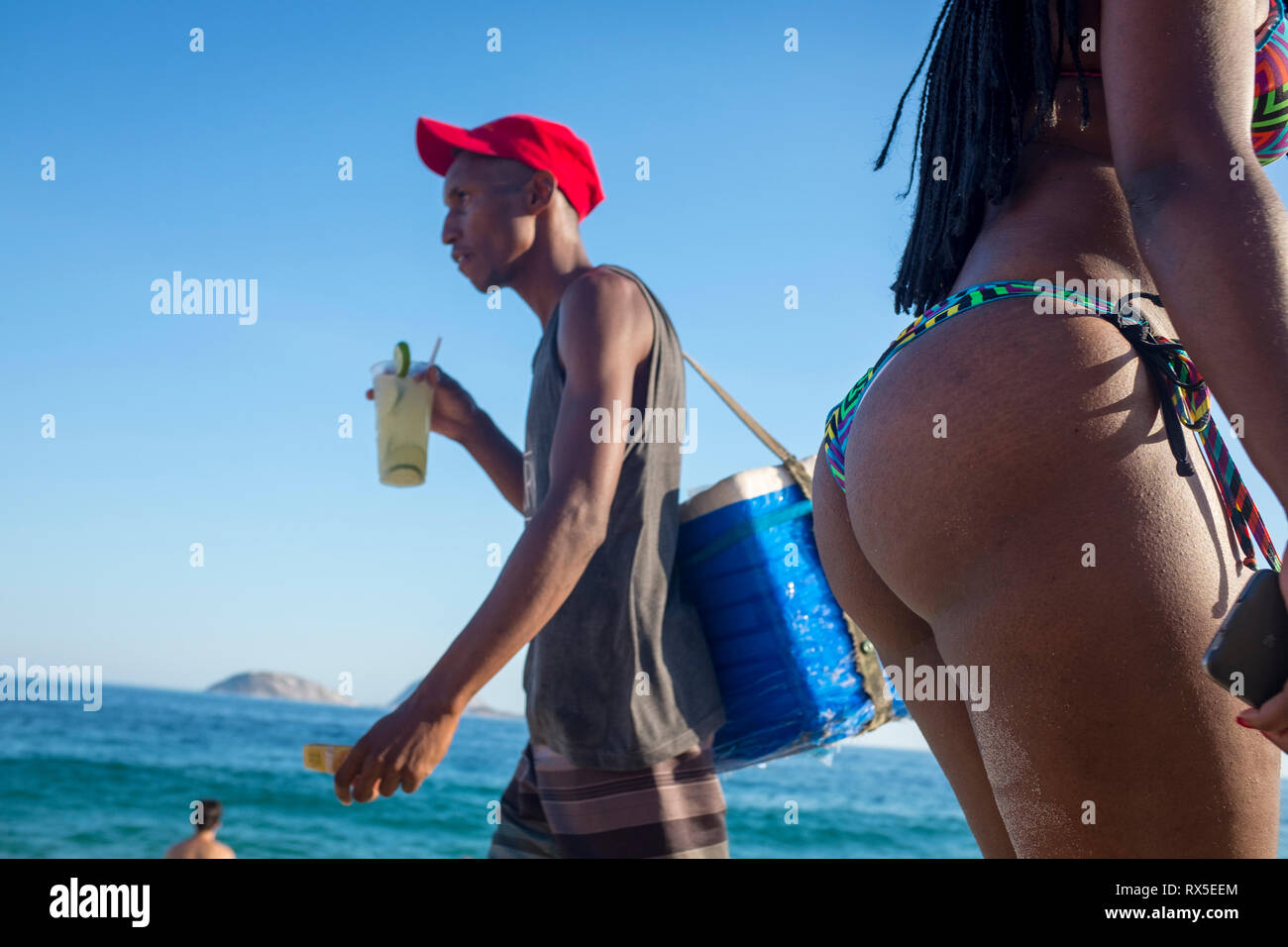 RIO DE JANEIRO - FEBRUARY 5, 2017: A beach vendor drinks a caipirinha cocktail as he passes a beachgoers in bikinis on the shore of Ipanema Beach. Stock Photo