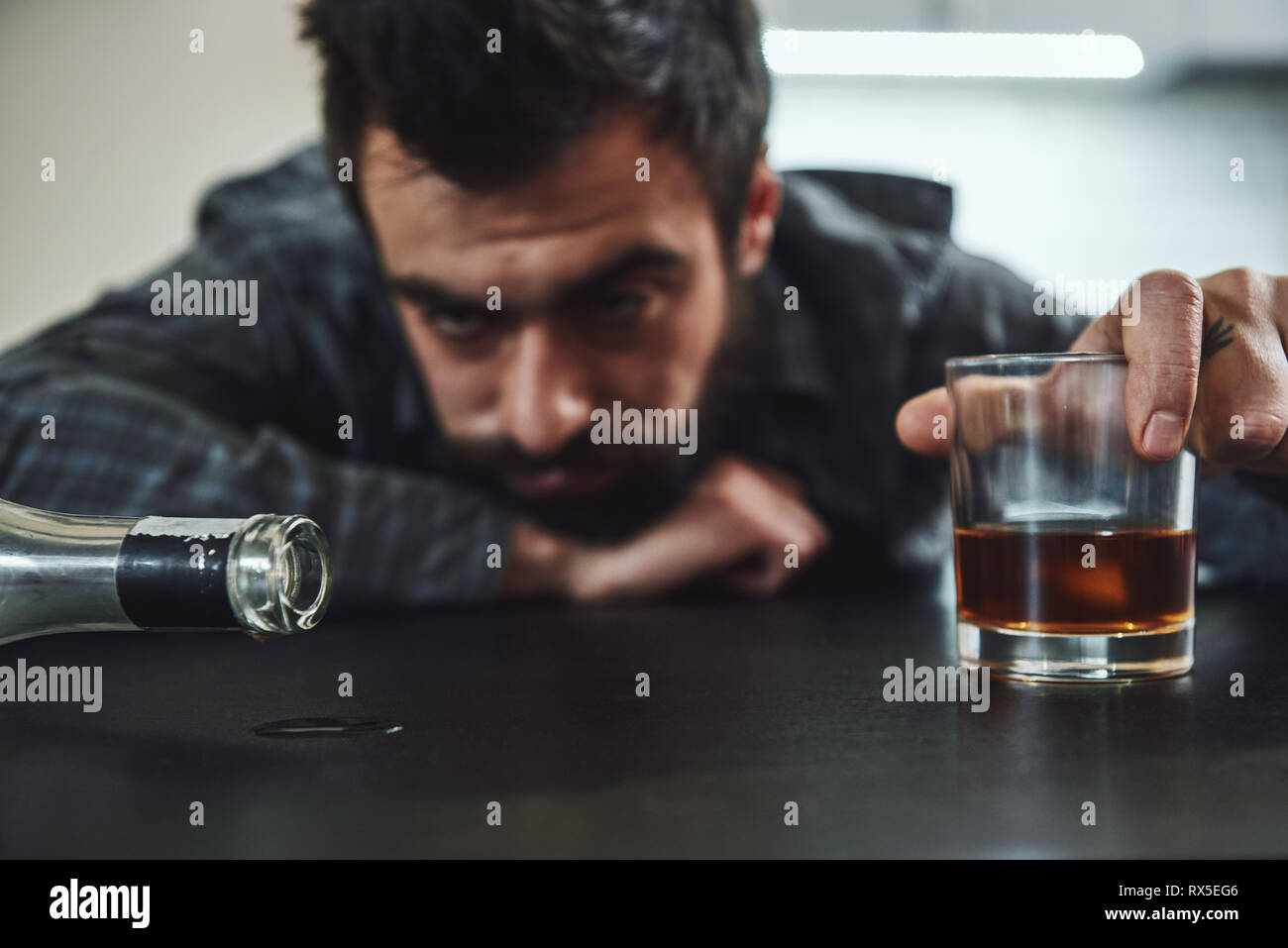 Dark-haired, sad and wasted alcoholic man lying on a table, in the kitchen. He holds a glass of whiskey while looking at empty bottle. Looking depress Stock Photo