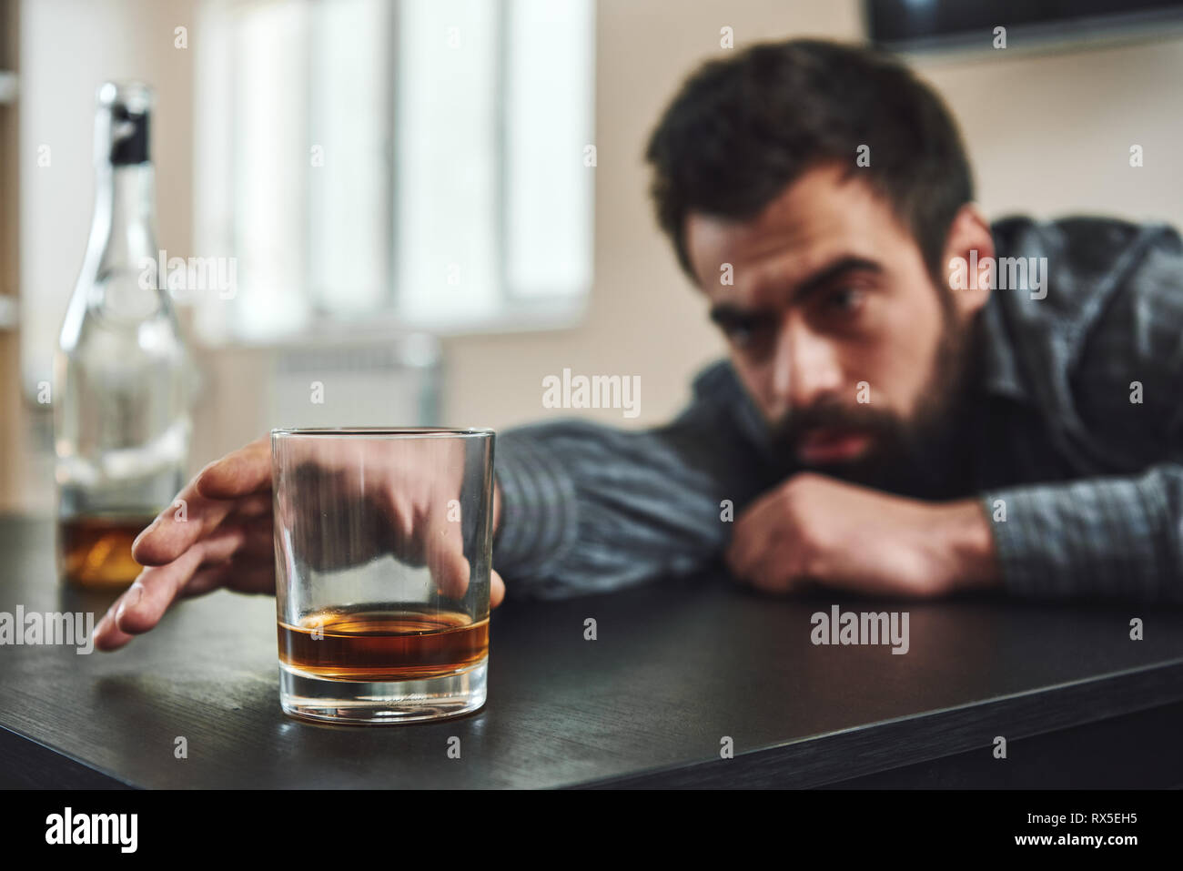 Dark-haired, sad and wasted alcoholic man lying on a table, in the kitchen. He looks at a glass of whiskey and tries to reach it. Looking depressed, l Stock Photo