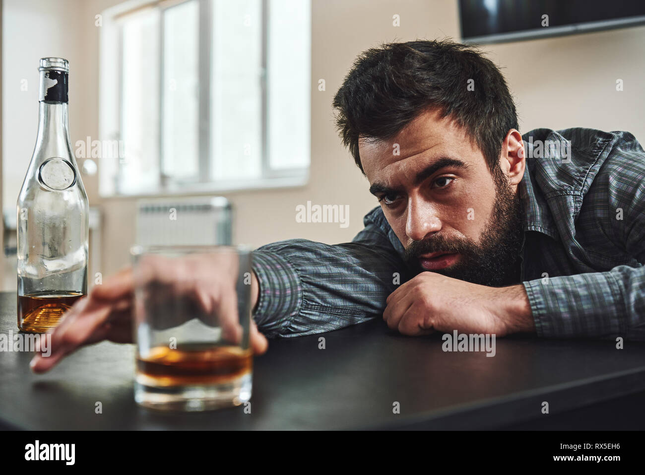Dark-haired, sad and wasted alcoholic man lying on a table, in the kitchen. He looks at a glass of whiskey and tries to reach it. Looking depressed, l Stock Photo