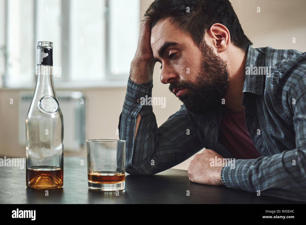 Drinking trouble. Depressed man sits at the table, holding hand on his head. A bottle and a glass of whiskey stand in front of him. He is thinking abo Stock Photo