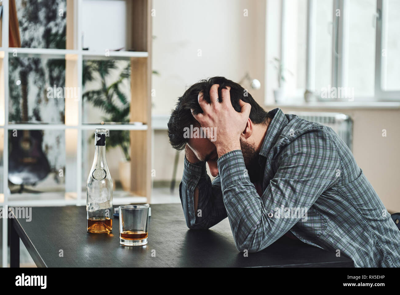 Drinking trouble. Depressed man sits at the table with his head in his hands. A bottle and a glass of whiskey stand in front of him. Side view Stock Photo