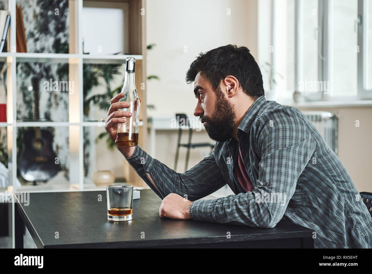 Drinking trouble. Depressed bearded man sits at the table with a bottle of whiskey in his hand. He looks at bottle while thinking about problems at wo Stock Photo