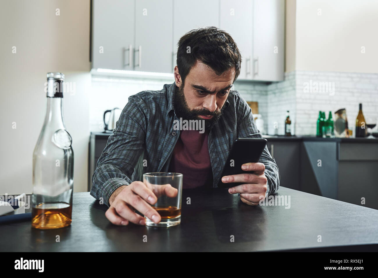 Portrait of young man in despair, sitting at table, drinking alcohol at home. He holds his phone while waiting for phone call. Male alcoholism concept Stock Photo
