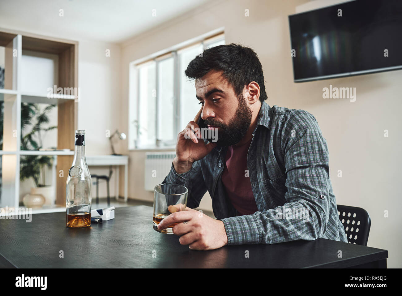 Portrait of young man in despair, sitting at table, drinking alcohol at home. He holds an empty glass while talking on the phone. Male alcoholism conc Stock Photo