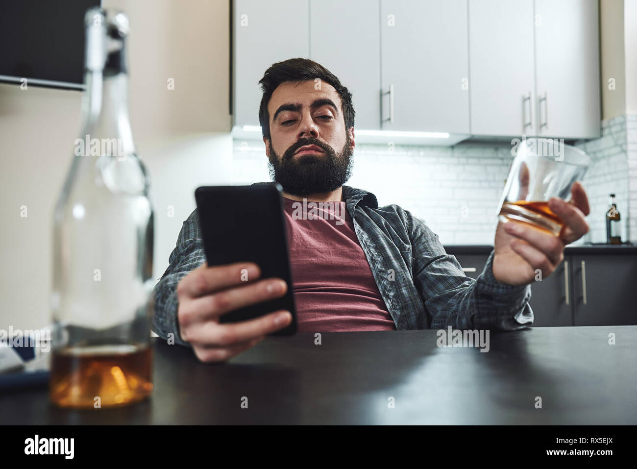 Portrait of young man in despair, sitting at table, drinking alcohol at home. He holds an empty glass while waiting for phone call. Male alcoholism co Stock Photo
