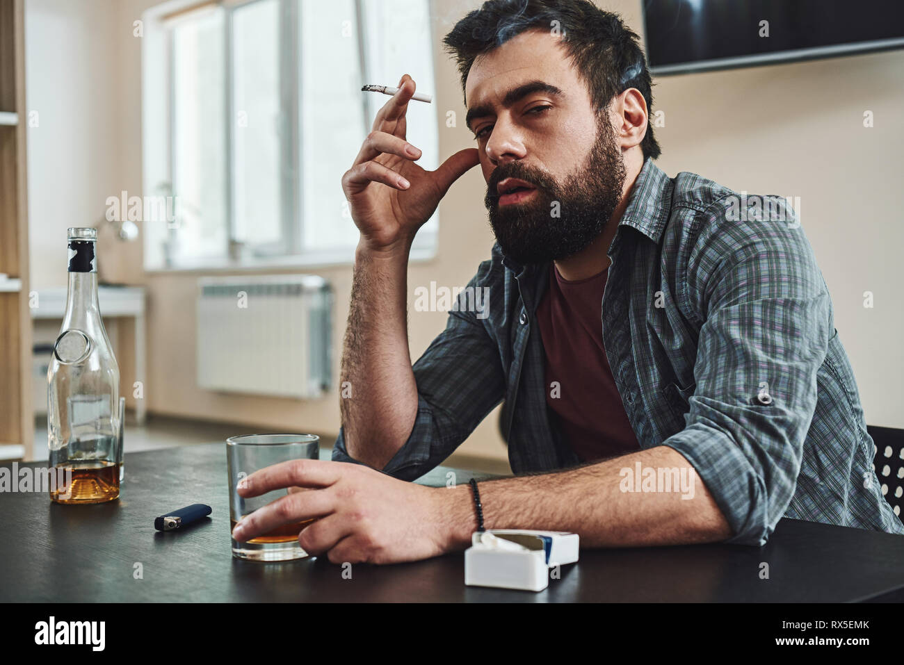 Portrait of dark-haired, sad and wasted alcoholic man sitting at the table, in the kitchen, smoking and drinking whiskey, holding glass and cigarette, Stock Photo