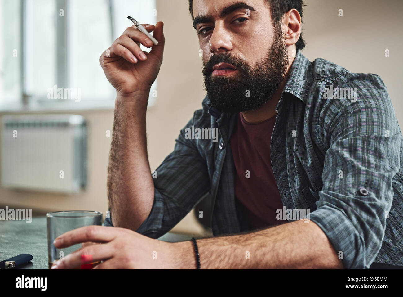 Cropped image of dark-haired, sad and wasted alcoholic man sitting at the table, in the kitchen, smoking and drinking whiskey, holding glass and cigar Stock Photo