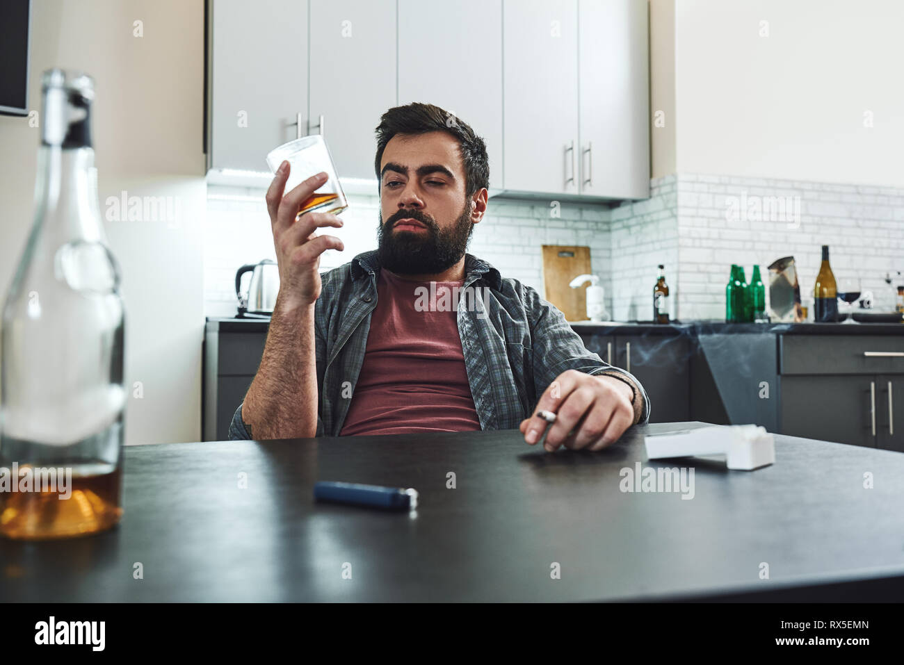 Dark-haired, sad and wasted alcoholic man sitting at the table, in the kitchen, smoking and drinking whiskey, holding glass and cigarette, completely  Stock Photo