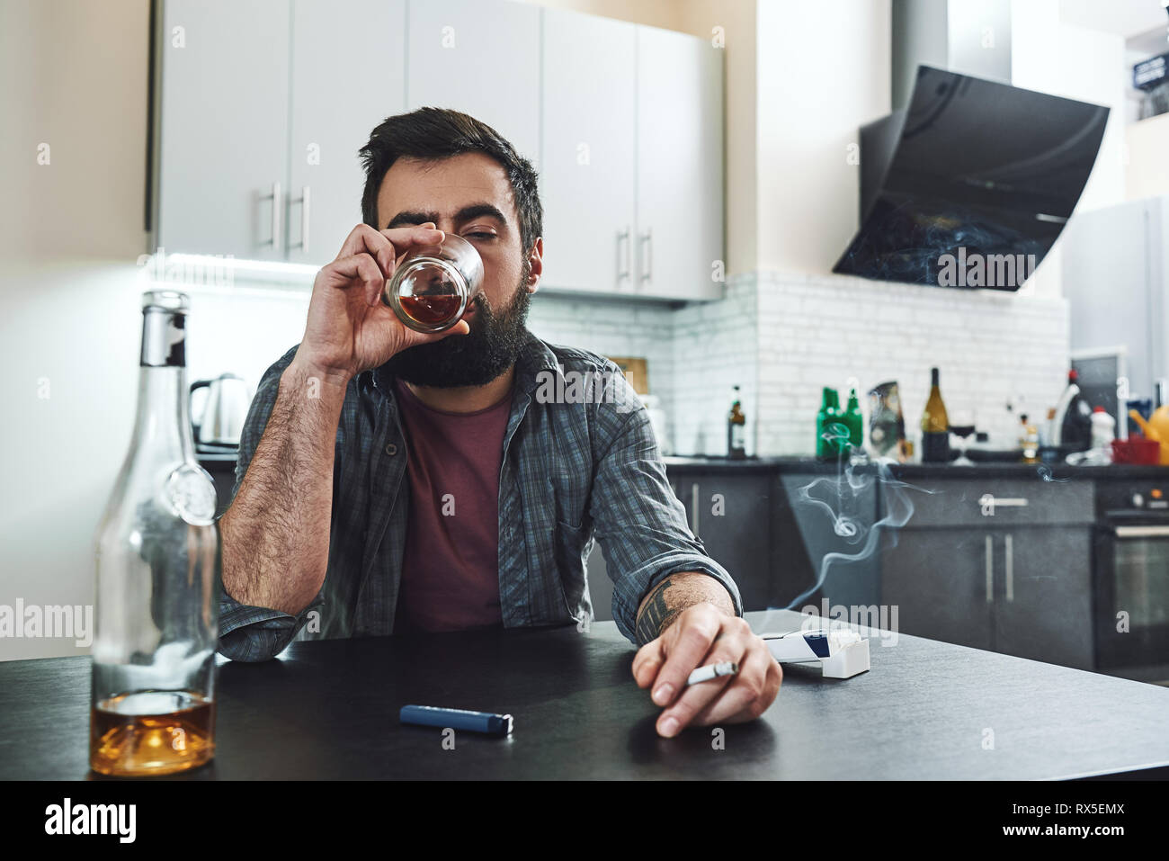 Dark-haired, sad and wasted alcoholic man sitting at the table, in the kitchen, smoking and drinking whiskey, holding glass, completely drunk, looking Stock Photo