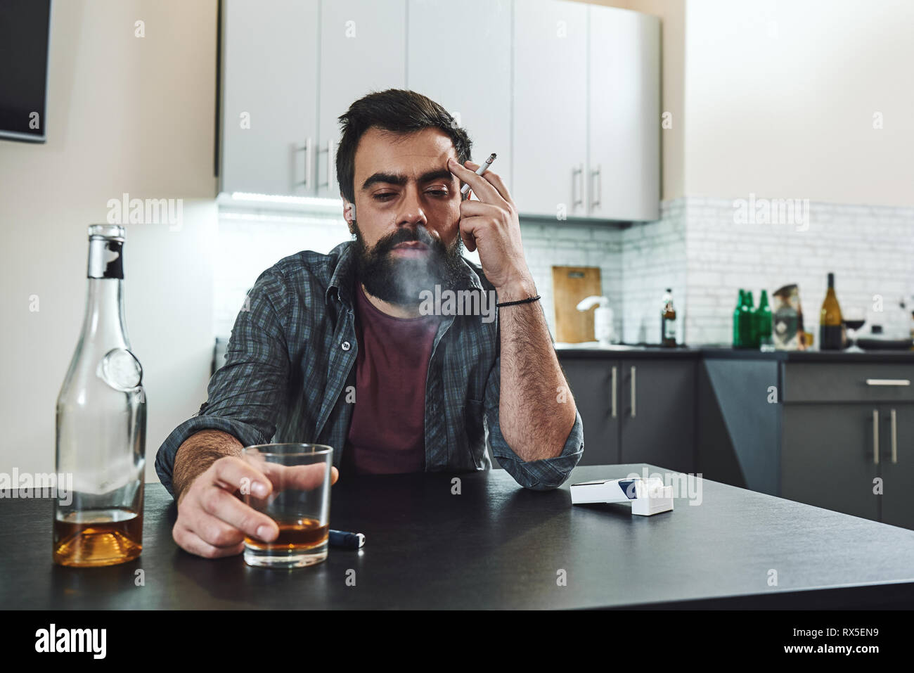 Dark-haired, sad and wasted alcoholic man sitting at the table, in the kitchen, smoking and drinking whiskey, holding glass, completely drunk, looking Stock Photo