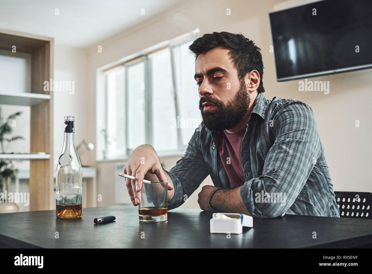 Dark-haired, sad and wasted alcoholic man sitting at the table, in the kitchen, drinking whiskey, holding glass, completely drunk, looking depressed,  Stock Photo