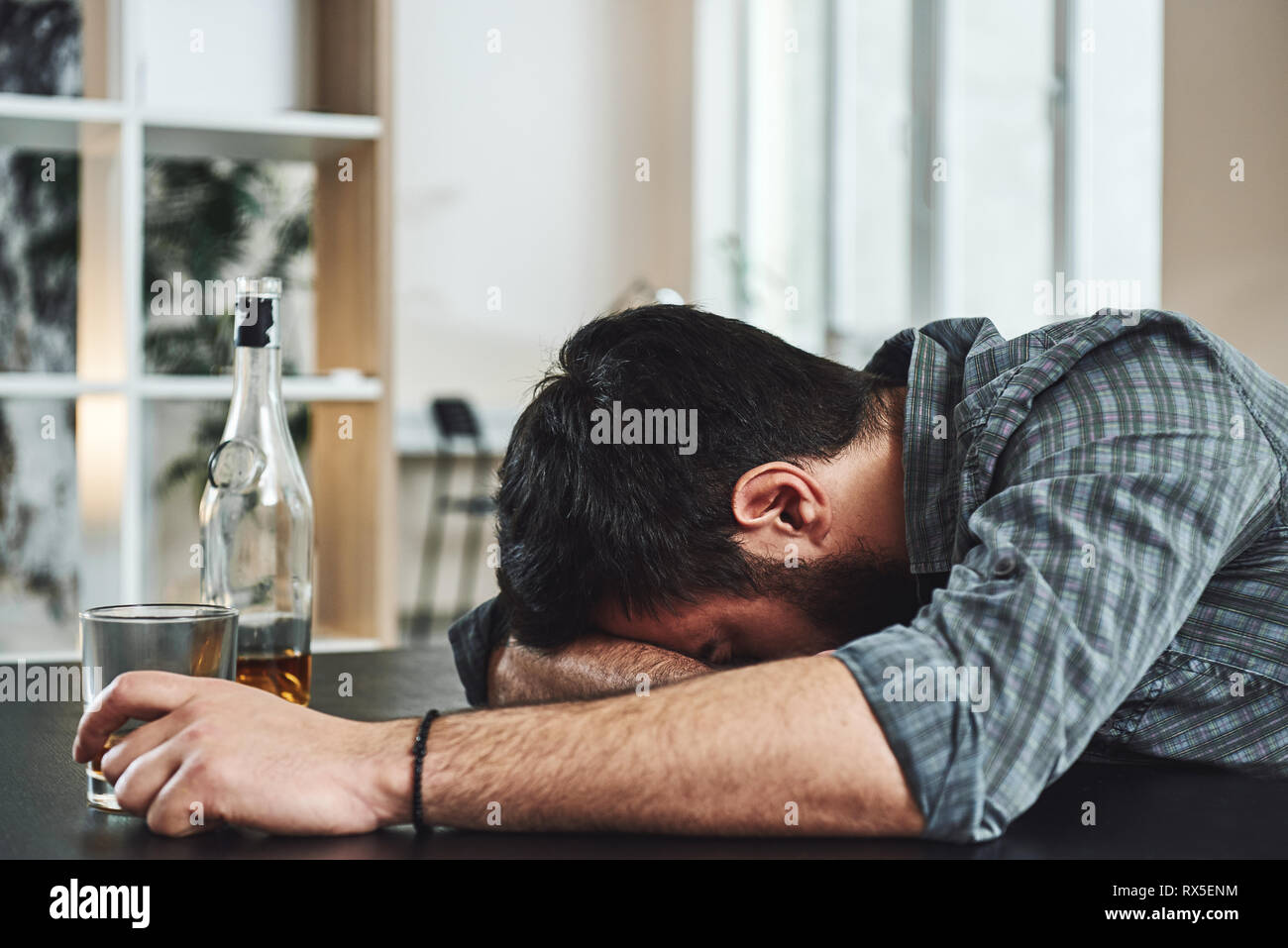 Dark-haired, sad and wasted alcoholic man lying on a table, in the kitchen, drinking whiskey, holding glass, completely drunk, looking depressed, lone Stock Photo