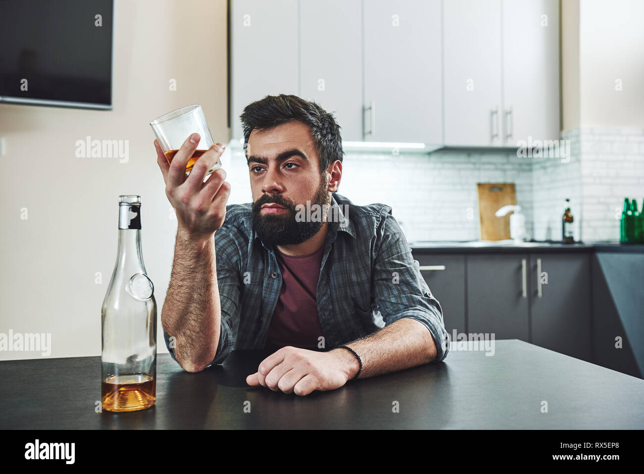 Drinking trouble. Depressed bearded man sits at the table with a glass of whiskey in his hand. He is thinking about problems at work and troubles in r Stock Photo