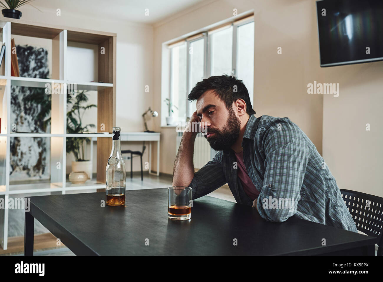 Drinking trouble. Depressed man sits at the table, holding hand on his head. A glass of whiskey stands in front of him. He is thinking about problems  Stock Photo