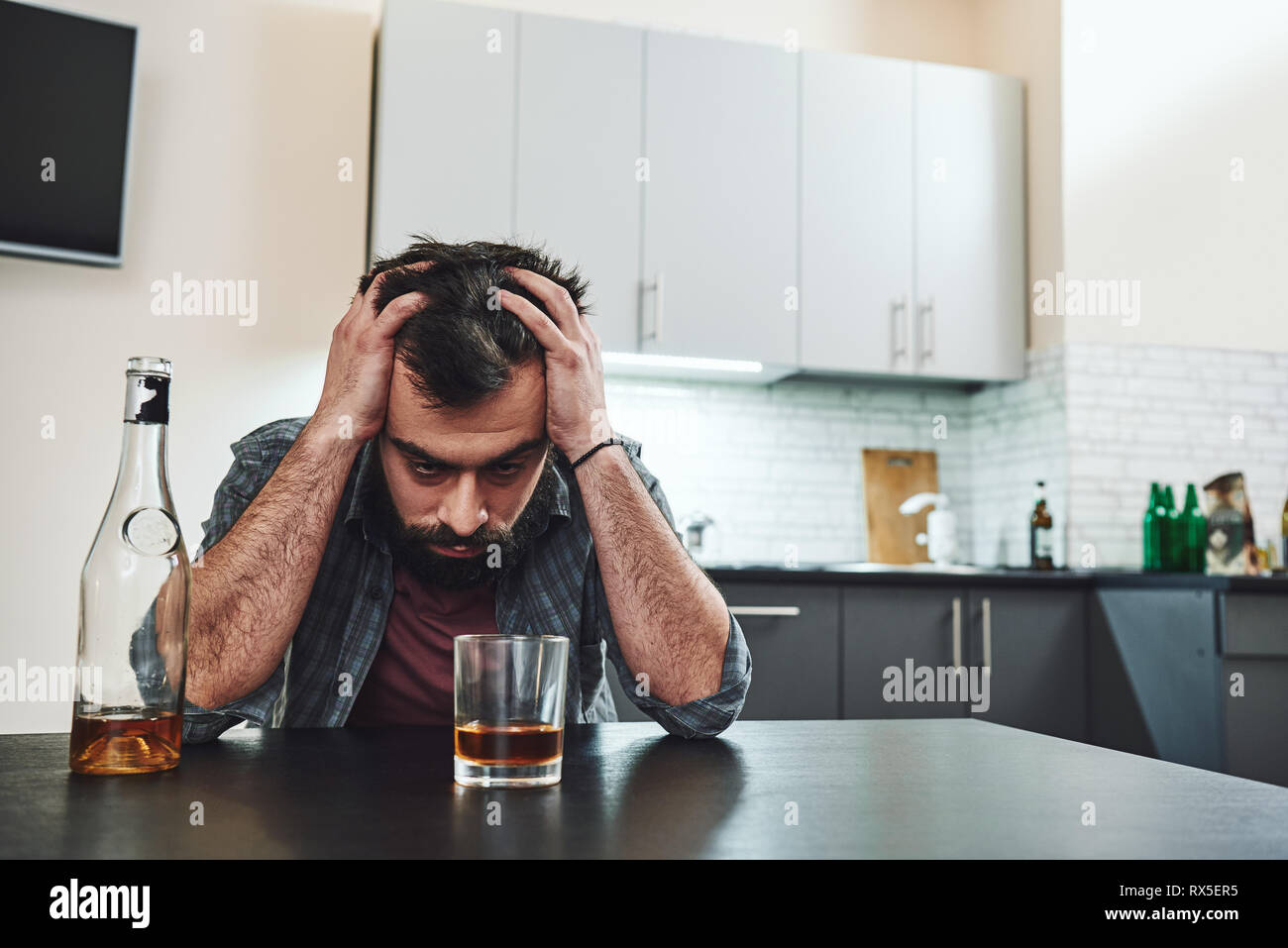 Drinking trouble. Depressed man sits at the table with his head in his hands. A glass of whiskey stands in front of him. Stock Photo