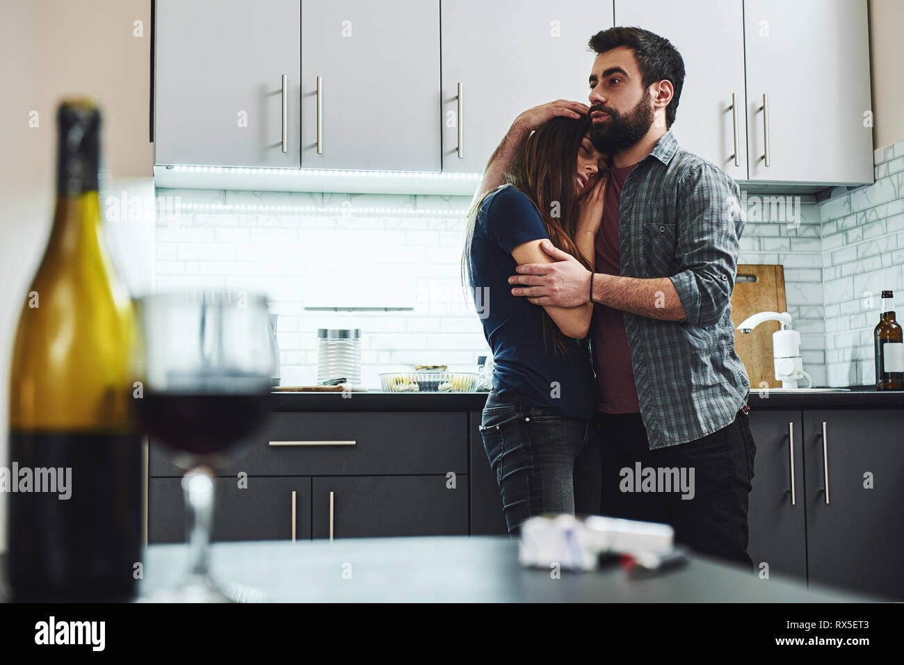 Couple of drunkards standing in the kitchen. Man hugging his drunk wife, she is almost sleeping. A bottle and a glass of red wine standing on the tabl Stock Photo