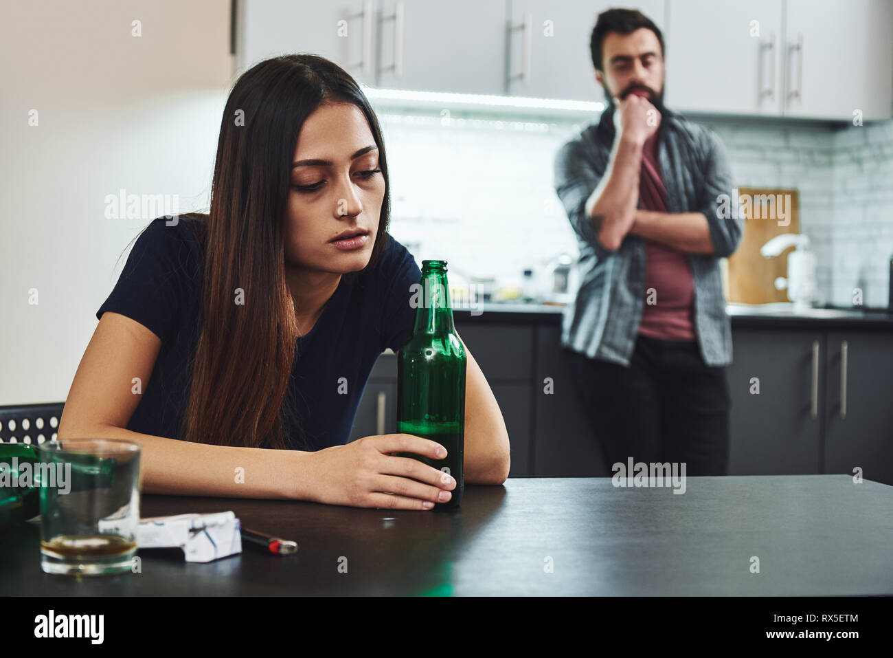 Drunkard. Angry man feeling despair while looking at his drunk wife. She is drinking in the kitchen, wasted. Empty bottles are on the table near her. Stock Photo