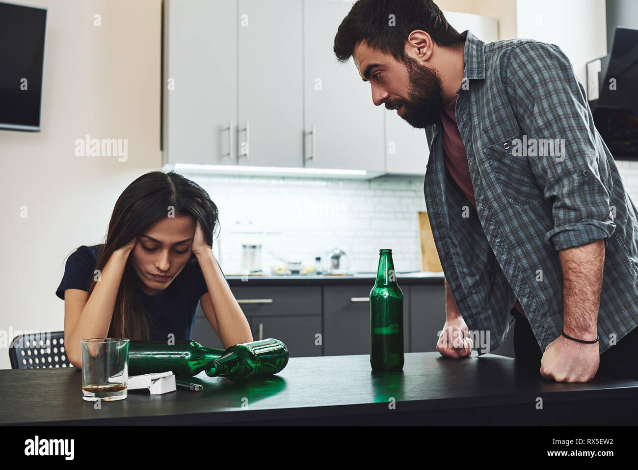 Drunkard. Angry man feeling despair while shouting at his drunk wife sitting in the kitchen. Empty bottles are on the table. Stock Photo