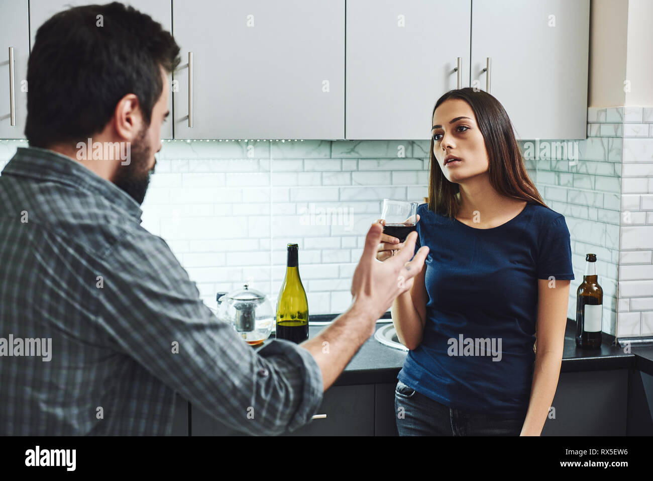 Drunkard. Dark-haired addicted woman arguing with her husband. She is standing in the kitchen with a glass of red wine looking at him and listening to Stock Photo